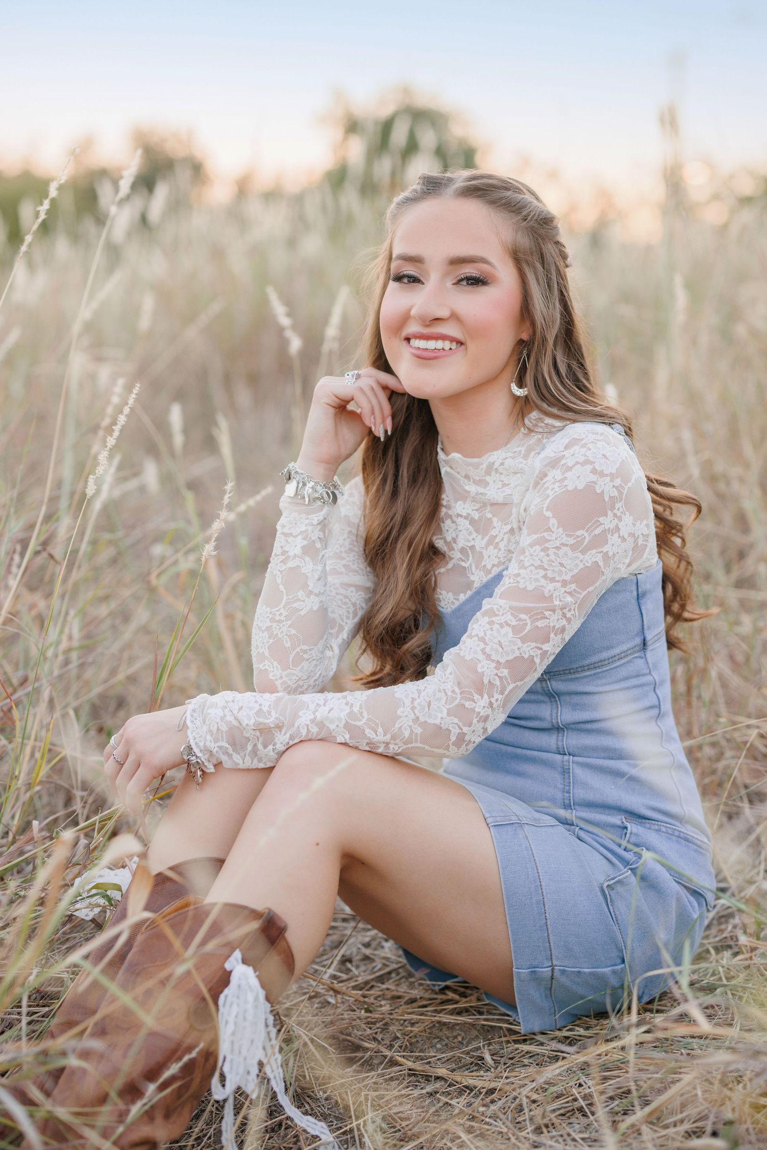 Portrait in a field at sunset wearing denim overalls and white lace top with long wavy hair and boots.