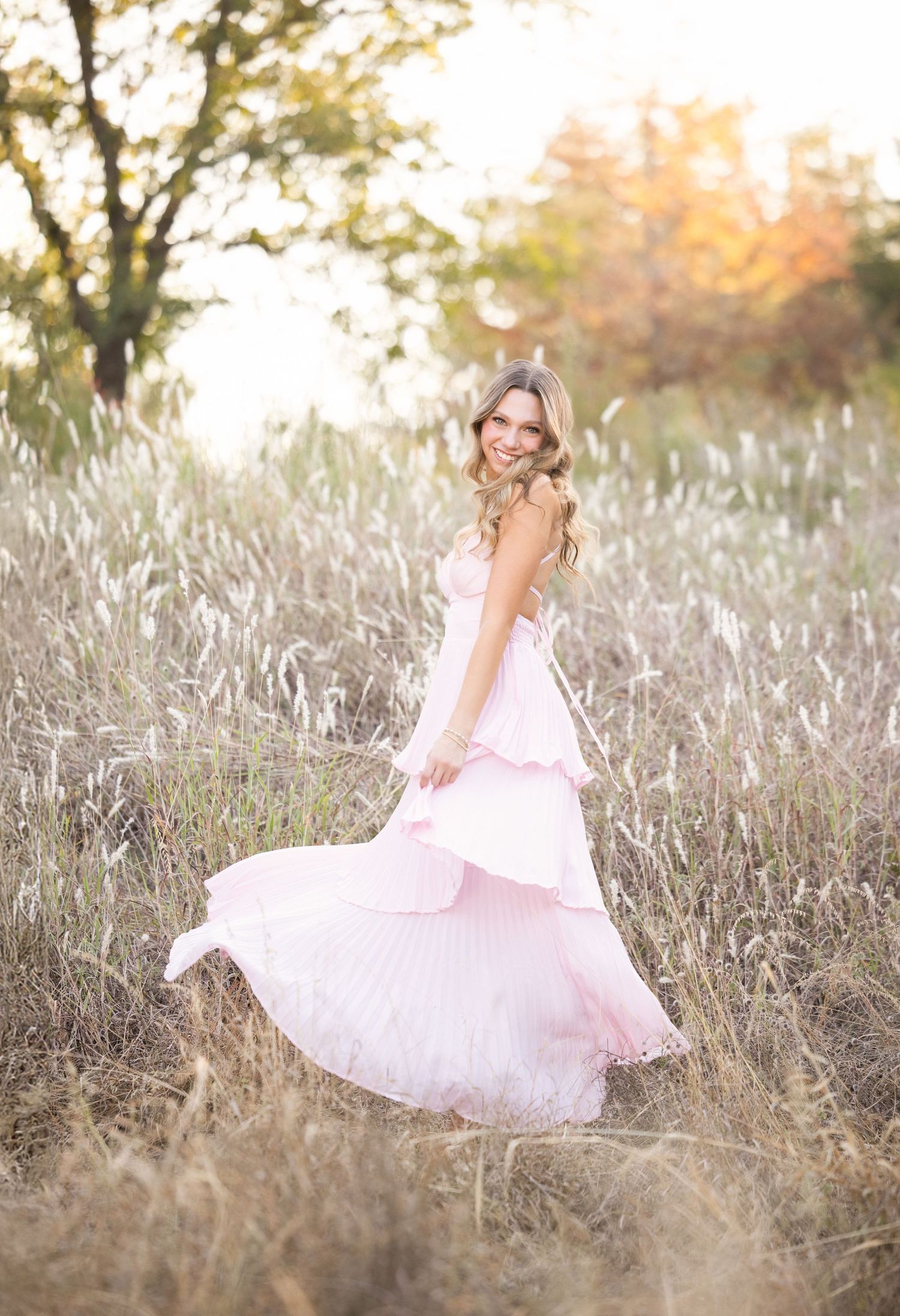 A pink flowing dress twirls gracefully in a sunlit field of tall autumn grass during golden hour.