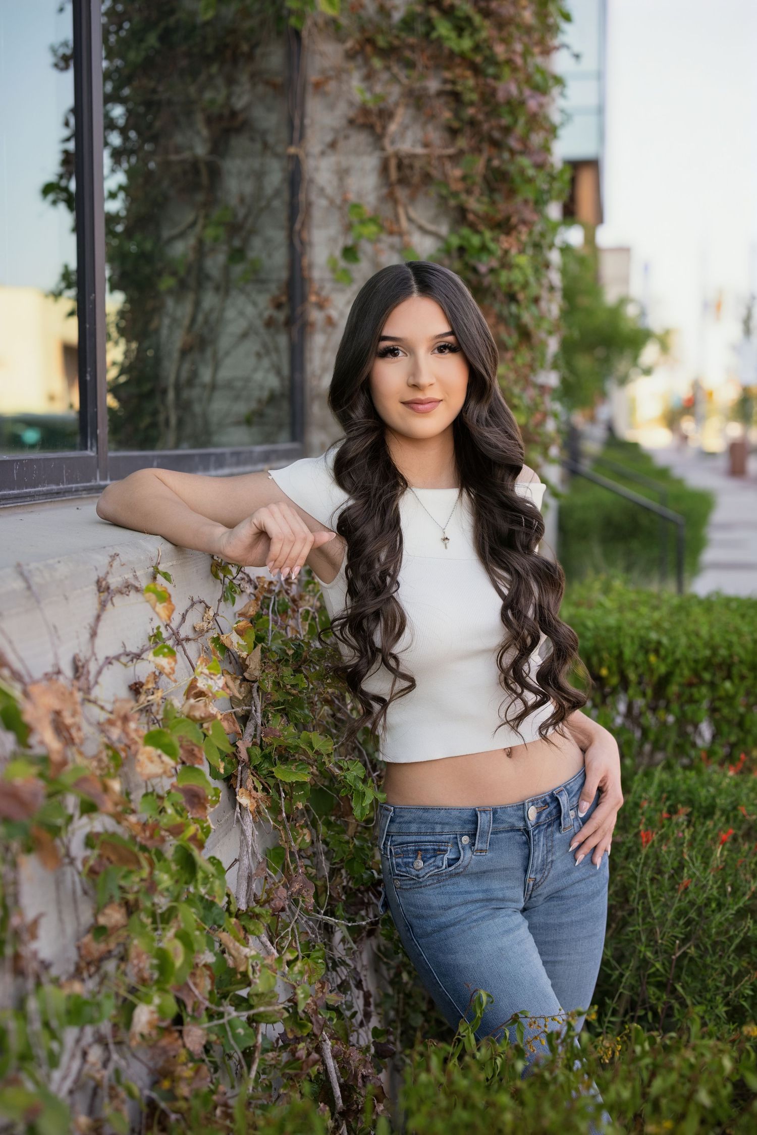 Young woman with long curly dark hair poses in white crop top and jeans beside ivy-covered pillar outdoors.