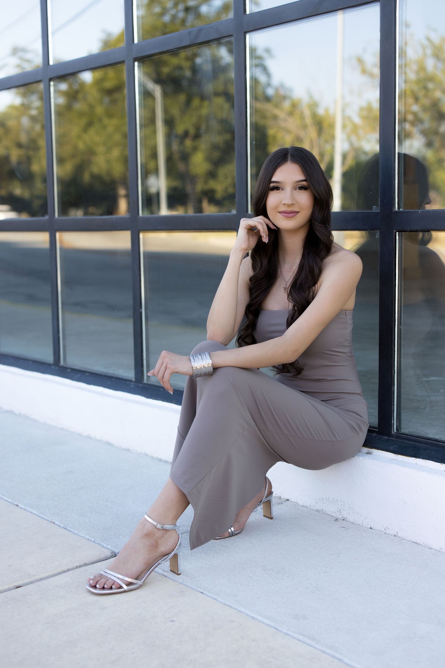 Young woman in gray jumpsuit and silver heels sitting gracefully against a modern black-framed window outdoors.