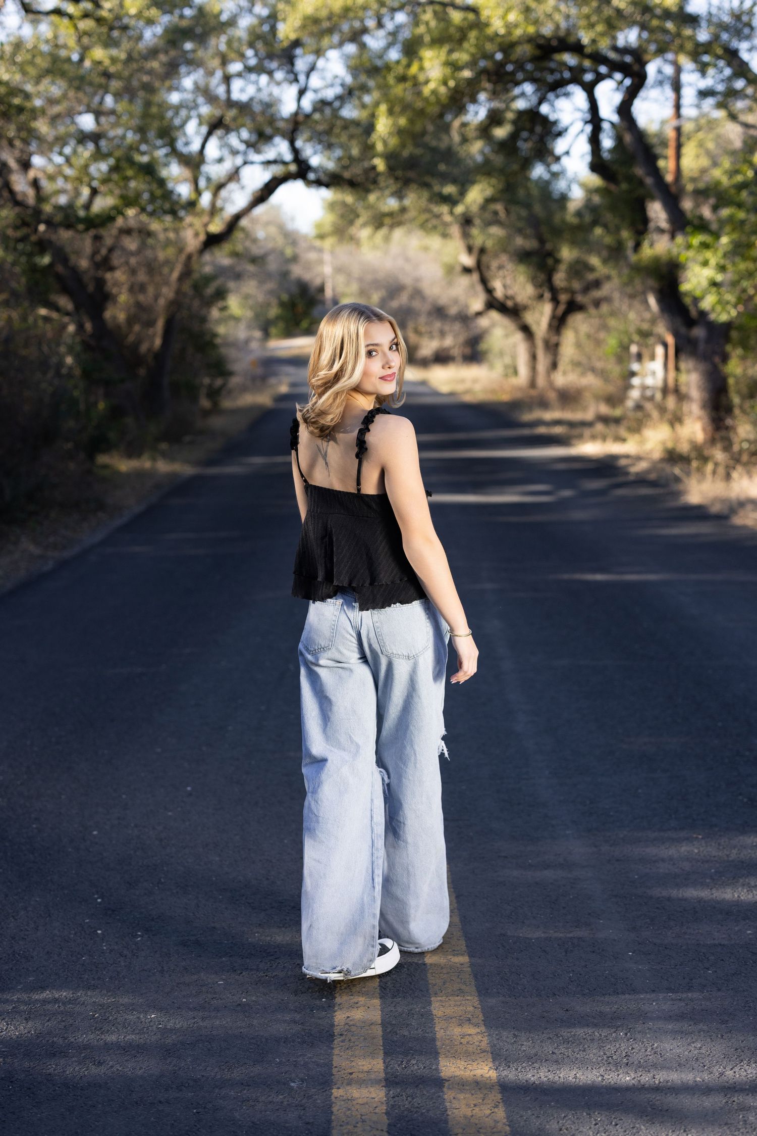 Young blonde woman in black tank top and wide-leg jeans standing on a tree-lined road, glancing back over her shoulder.