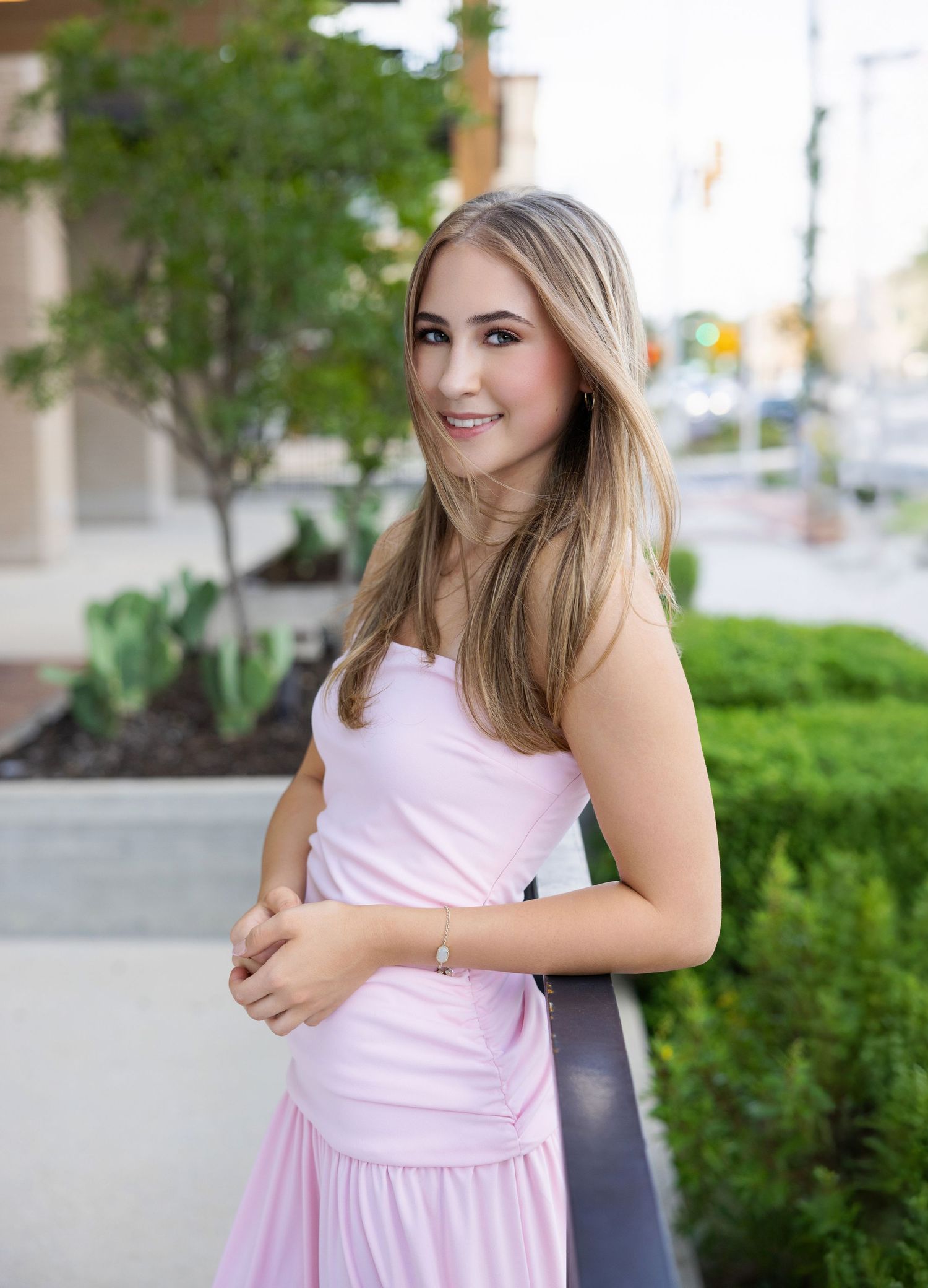 Young woman in a pink strapless dress smiling outdoors near greenery and urban buildings.
