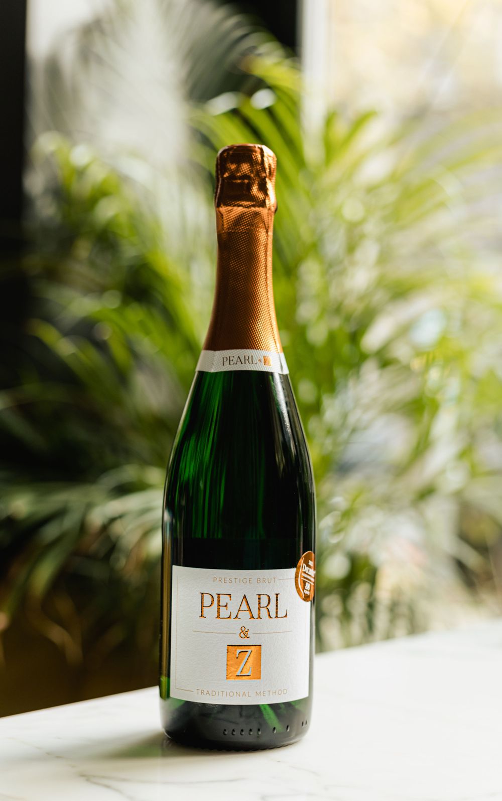 Pearl champagne bottle displayed on white surface with tropical plants in background.