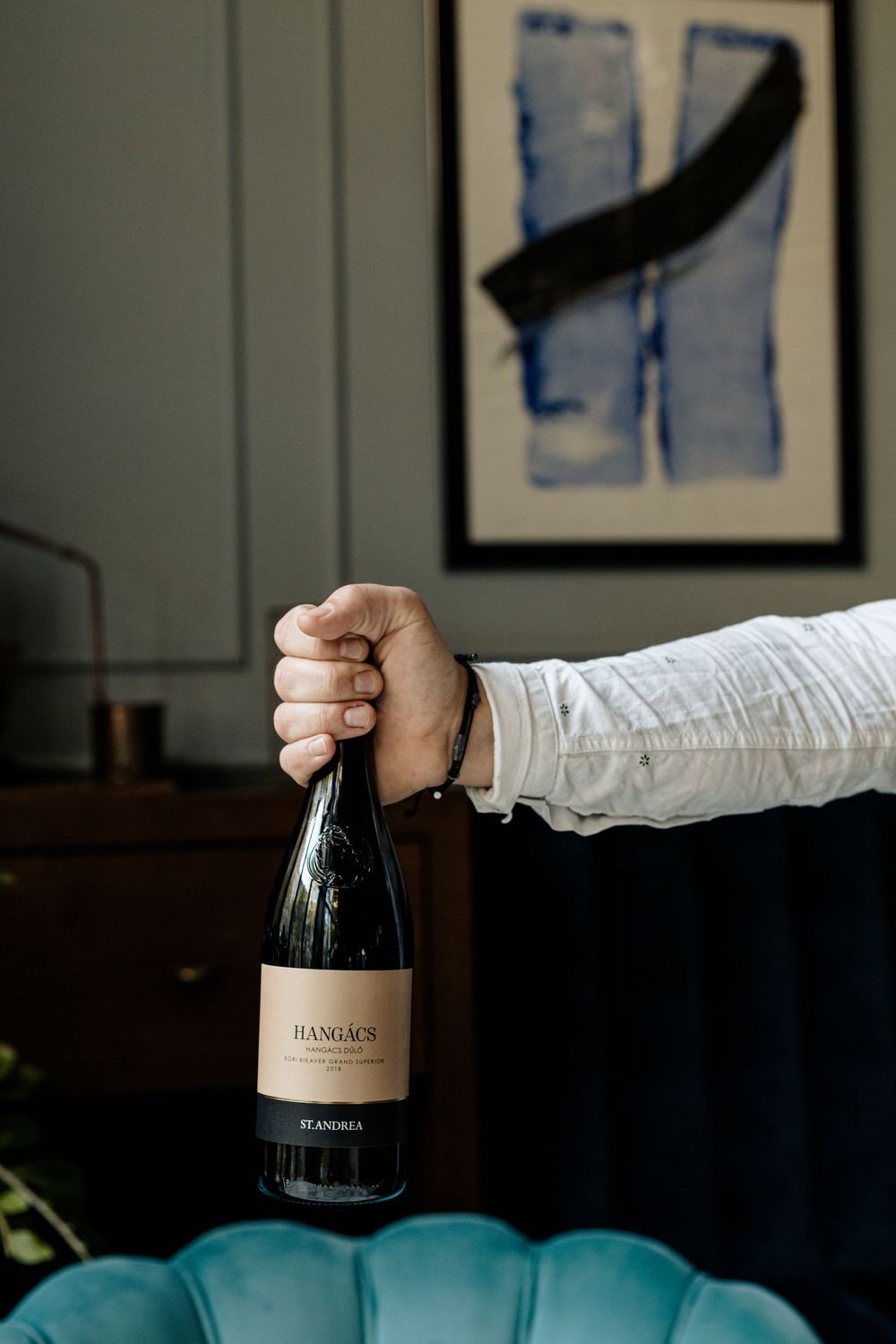 Hand holding wine bottle against blue velvet chair and modern wall art.