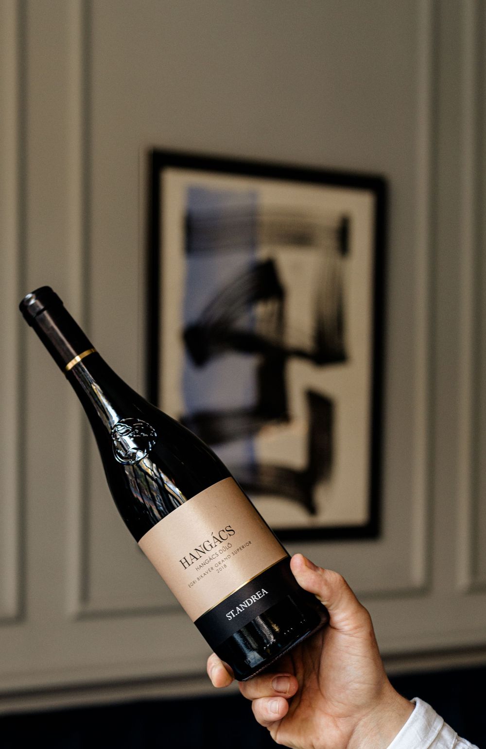 Hand holding wine bottle against wall with framed artwork in background.