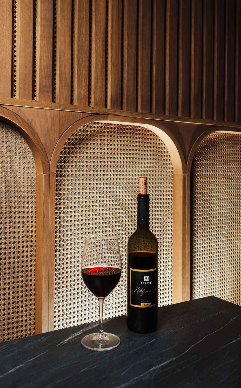 Red wine bottle and glass displayed in elegant wooden alcove with cane backing.