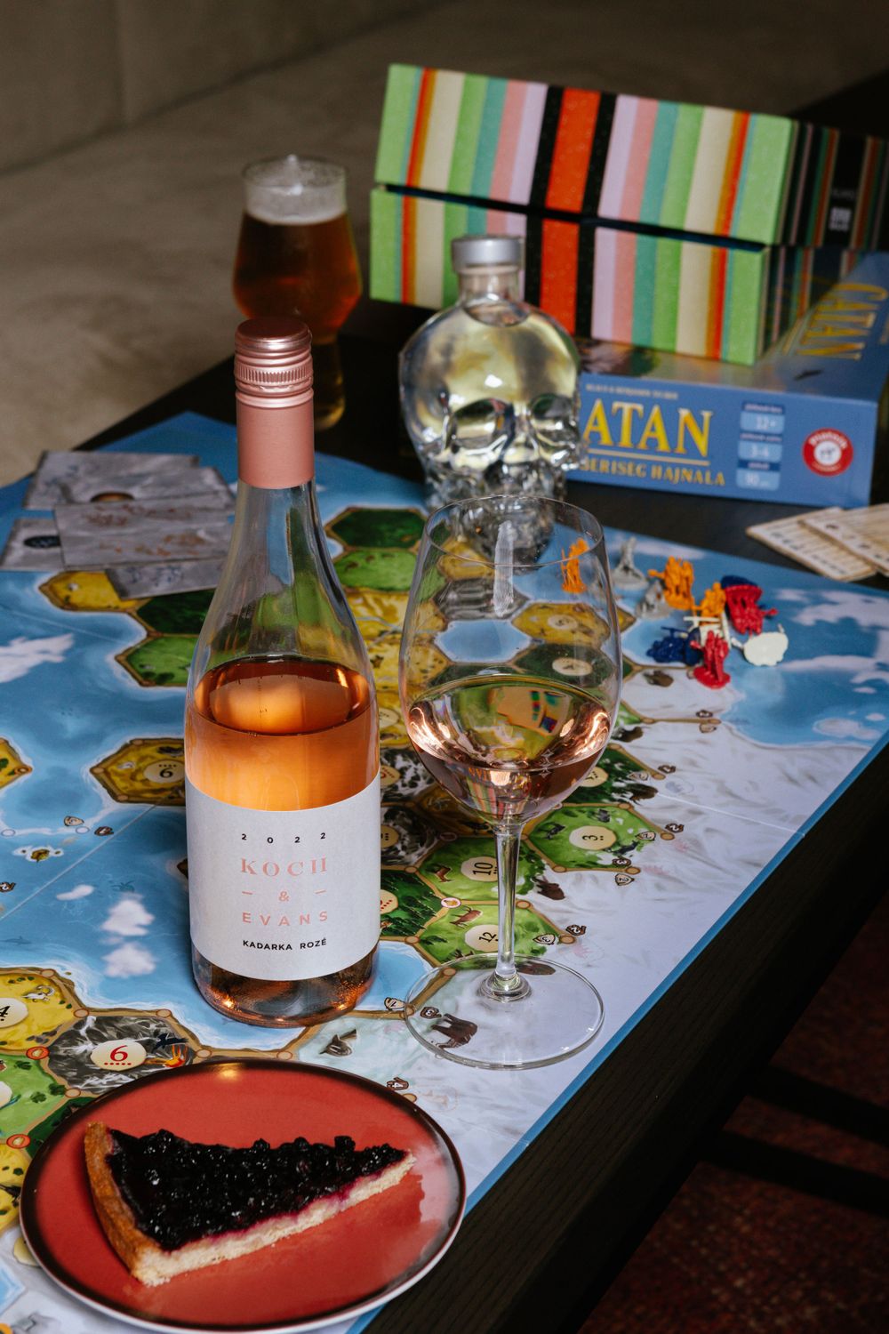 Board game setup with rosé wine, skull vodka bottle, and dessert on Catan game board.