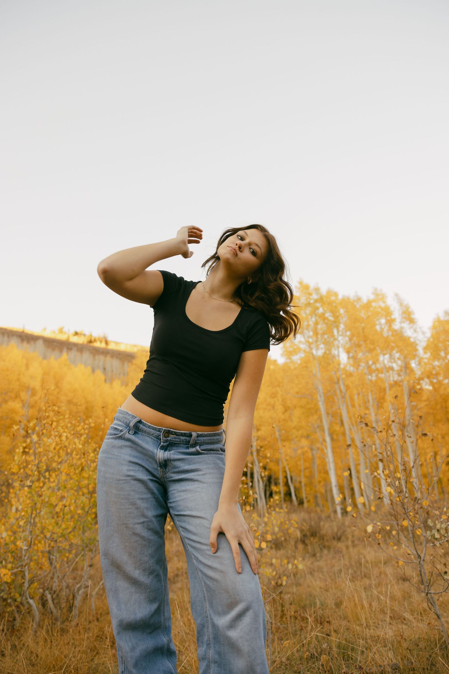 Person in black crop top and jeans poses against autumn landscape with golden foliage and white birch trees.