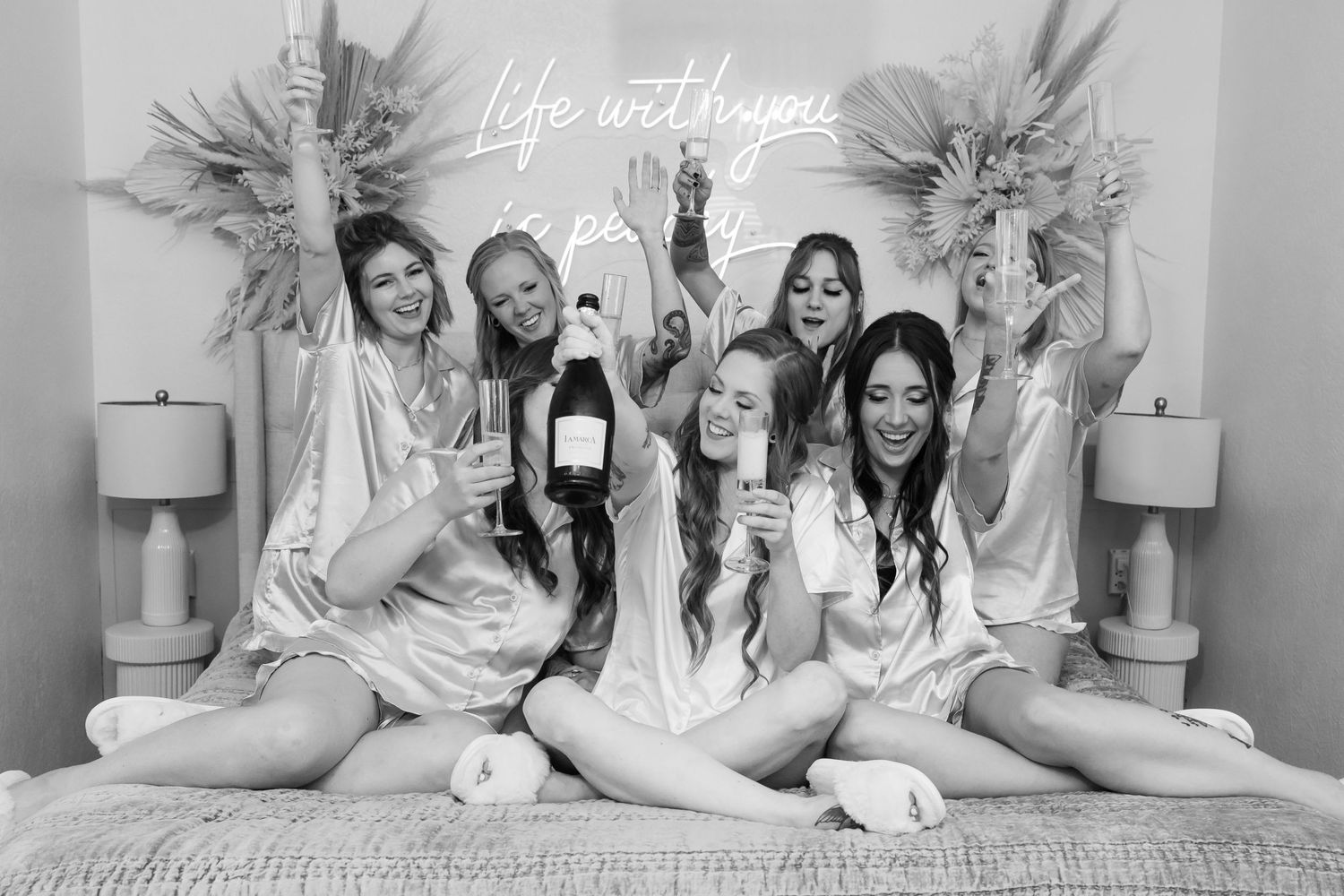 A group celebrates in matching white robes on a bed with champagne and neon signage.