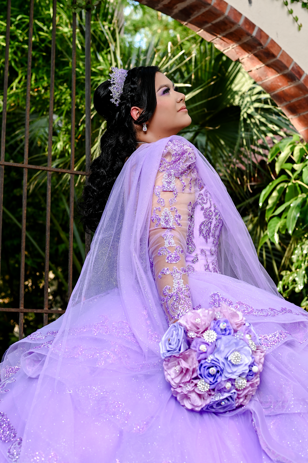 Elegant purple quinceanera gown with floral bouquet photographed in garden setting.