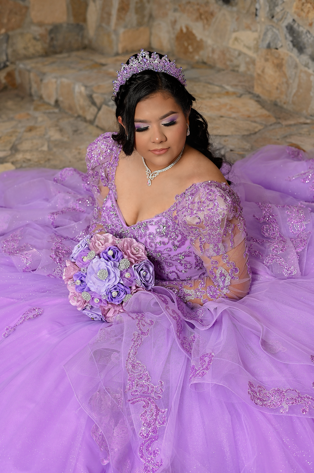 A stunning purple quinceañera ball gown with delicate lace details and matching tiara on stone background.