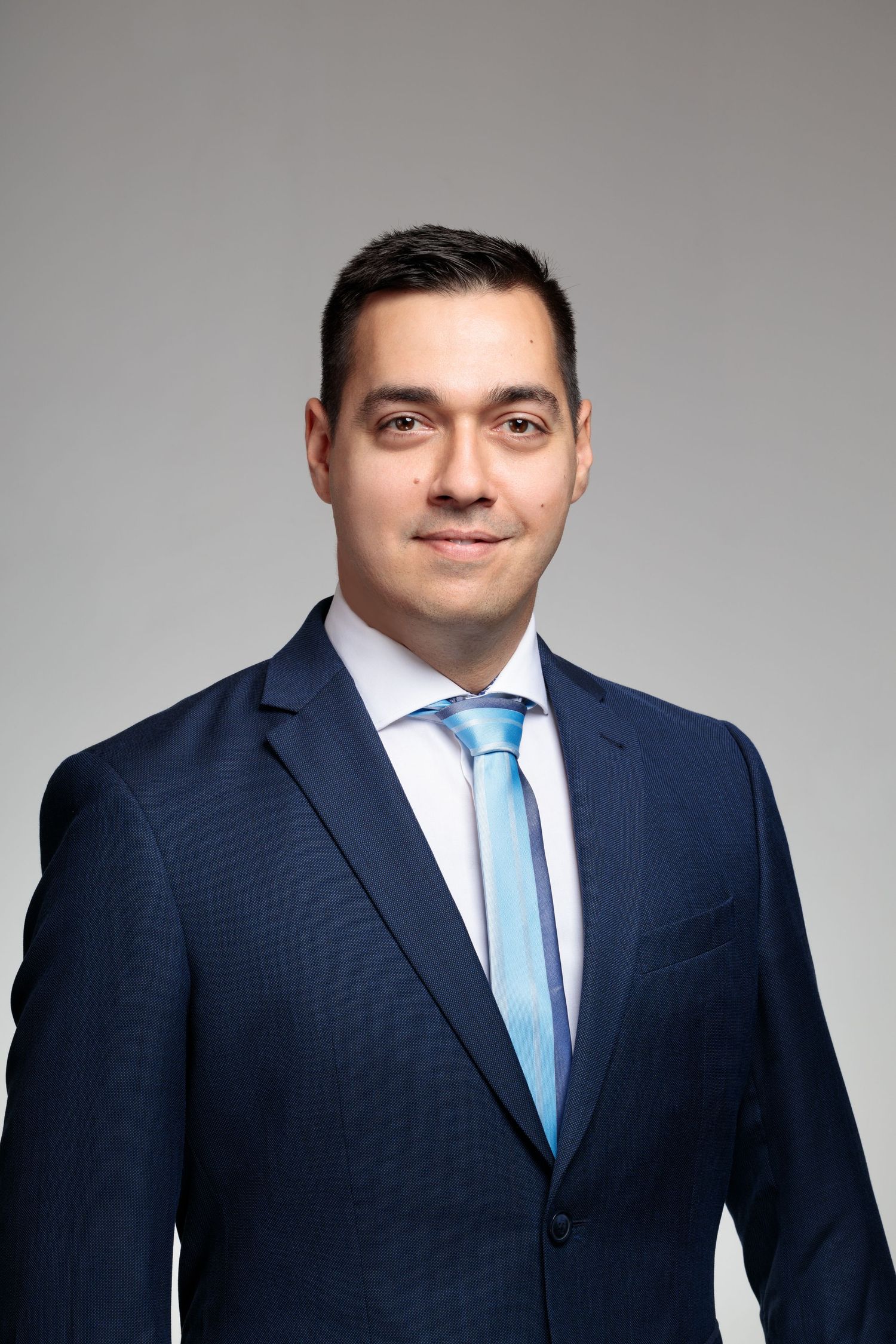 Professional portrait in navy suit with light blue tie against grey background.