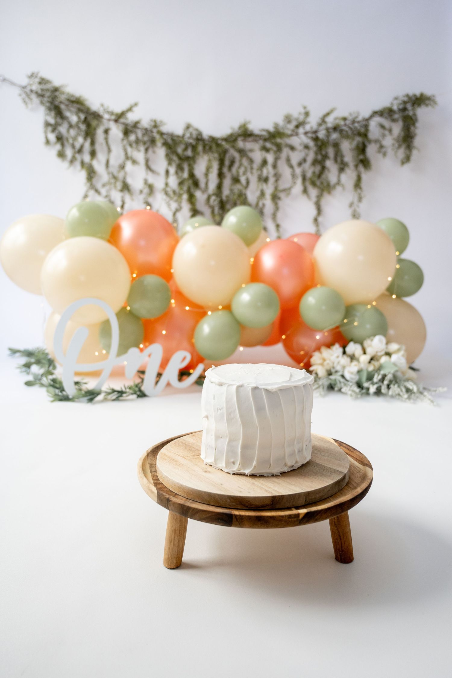 Wooden cake stand with white cake surrounded by pastel balloons and greenery garland.