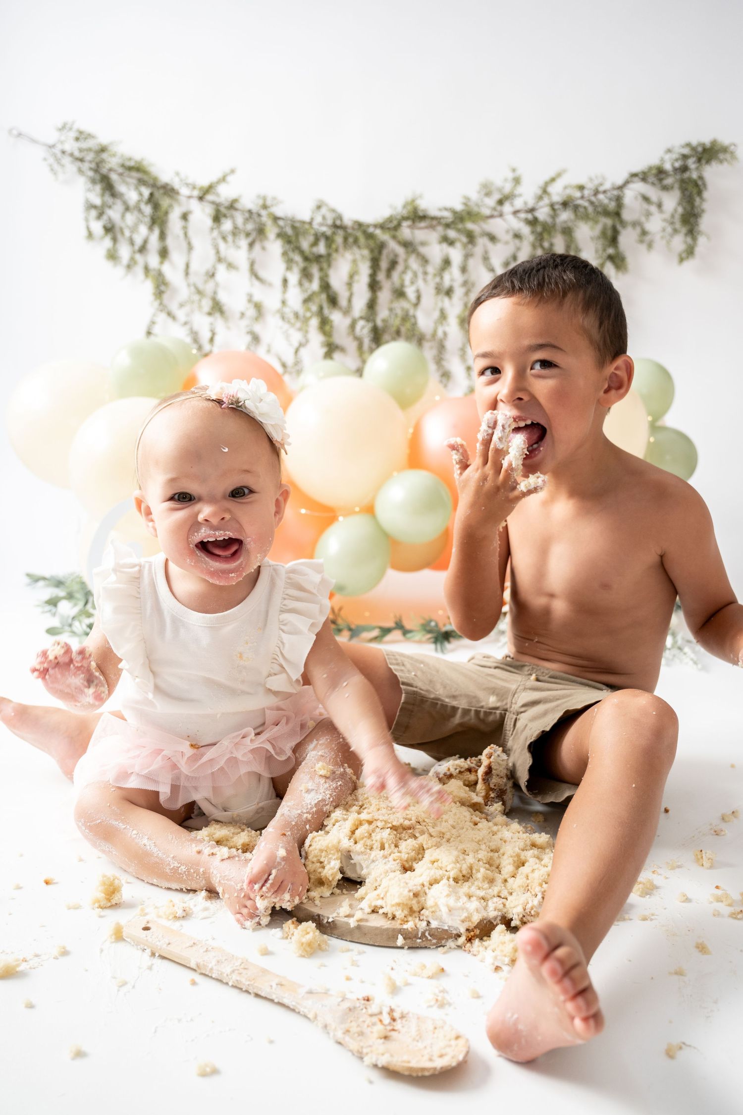 Two children laugh and play with cake during a festive cake smash photo session with pastel balloons and greenery.