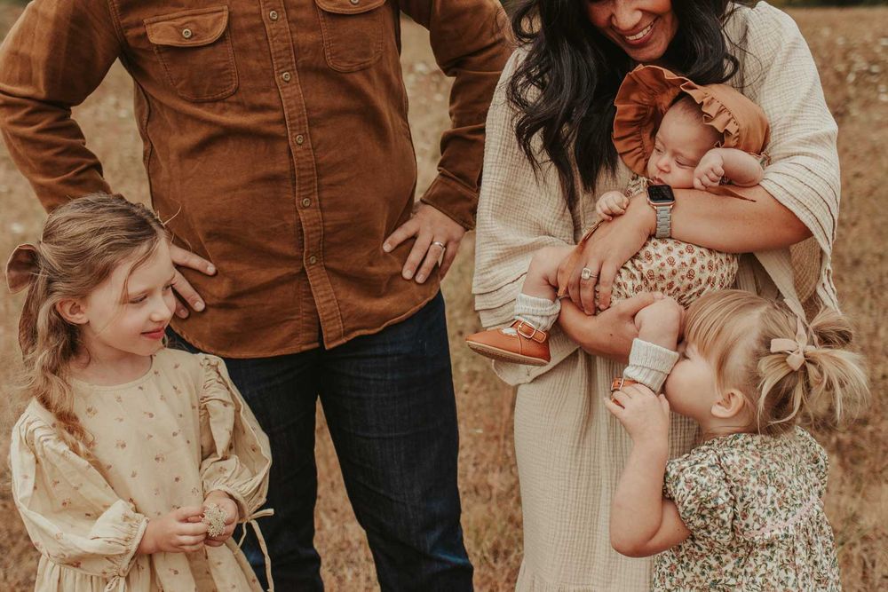 mom dad two little girls and one baby girls in an open field with rolling hills in the background tickling babies feet