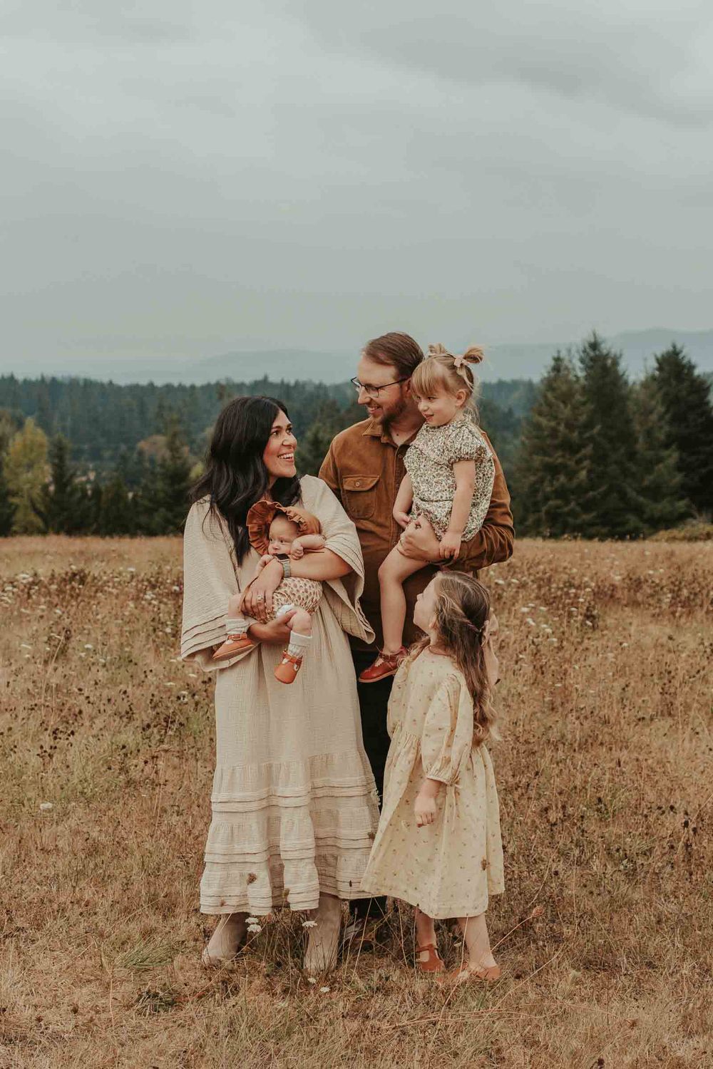 mom dad two little girls and one baby girls in an open field with rolling hills in the background smiling at each other