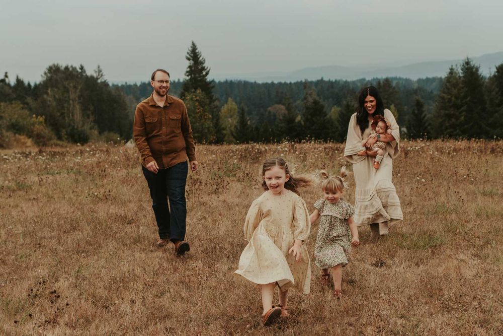 mom dad two little girls and one baby girls in an open field with rolling hills in the background smiling and playing