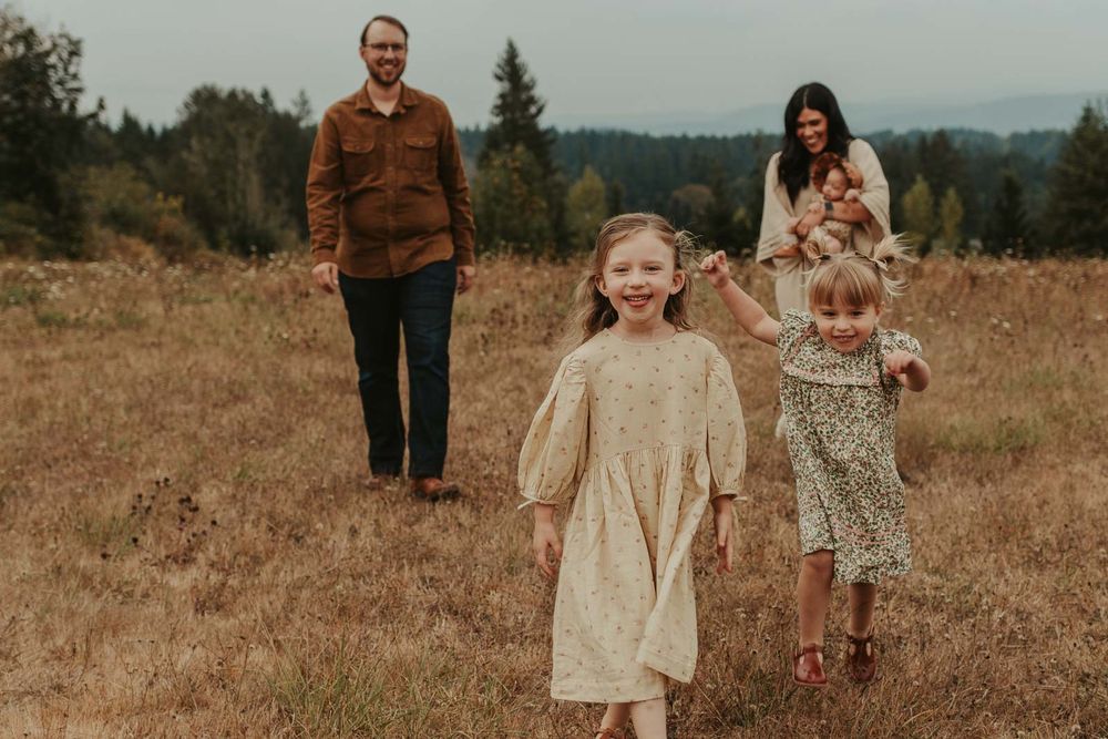 mom dad two little girls and one baby girls in an open field with rolling hills in the background smiling and playing