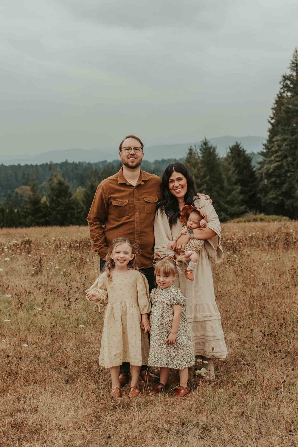 mom dad two little girls and one baby girls in an open field with rolling hills in the background smiling at the camera