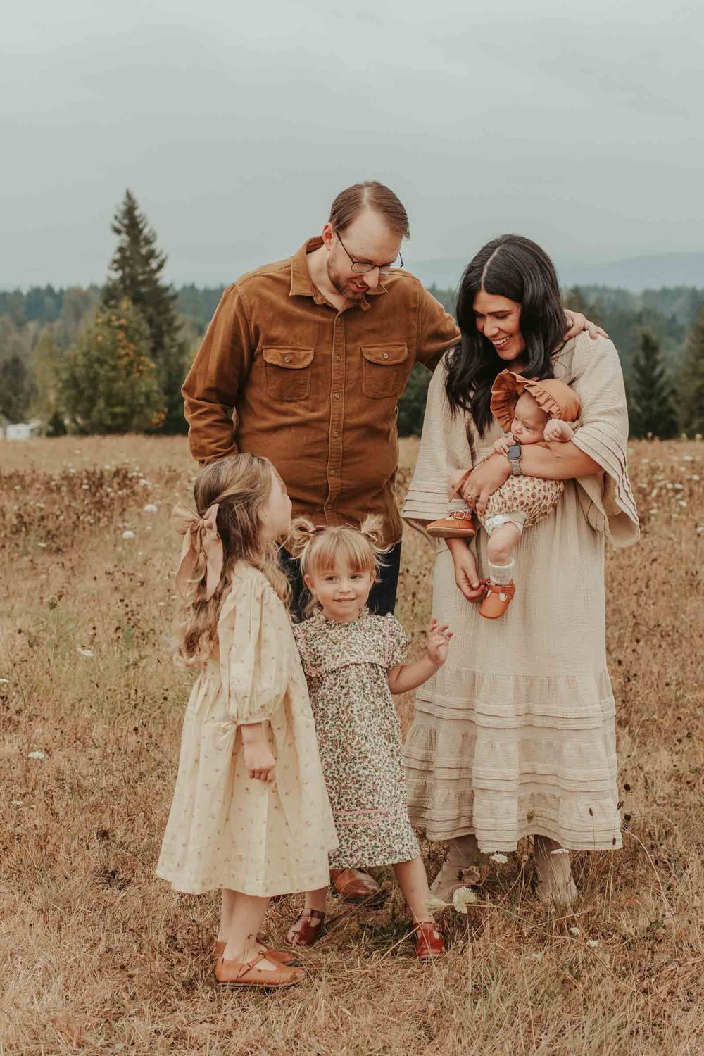 mom dad two little girls and one baby girls in an open field with rolling hills in the background smiling looking at mom