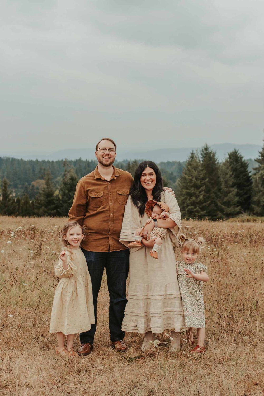 mom dad two little girls and one baby girls in an open field with rolling hills in the background smiling at the camera