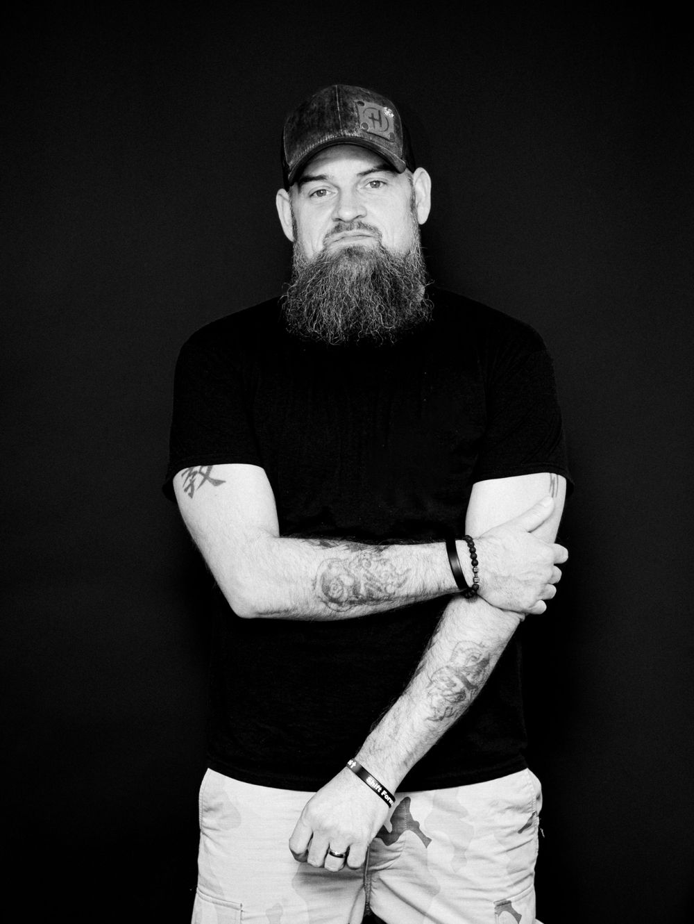 Black and white portrait showing person in baseball cap and dark shirt with long beard and tattoos against dark background.
