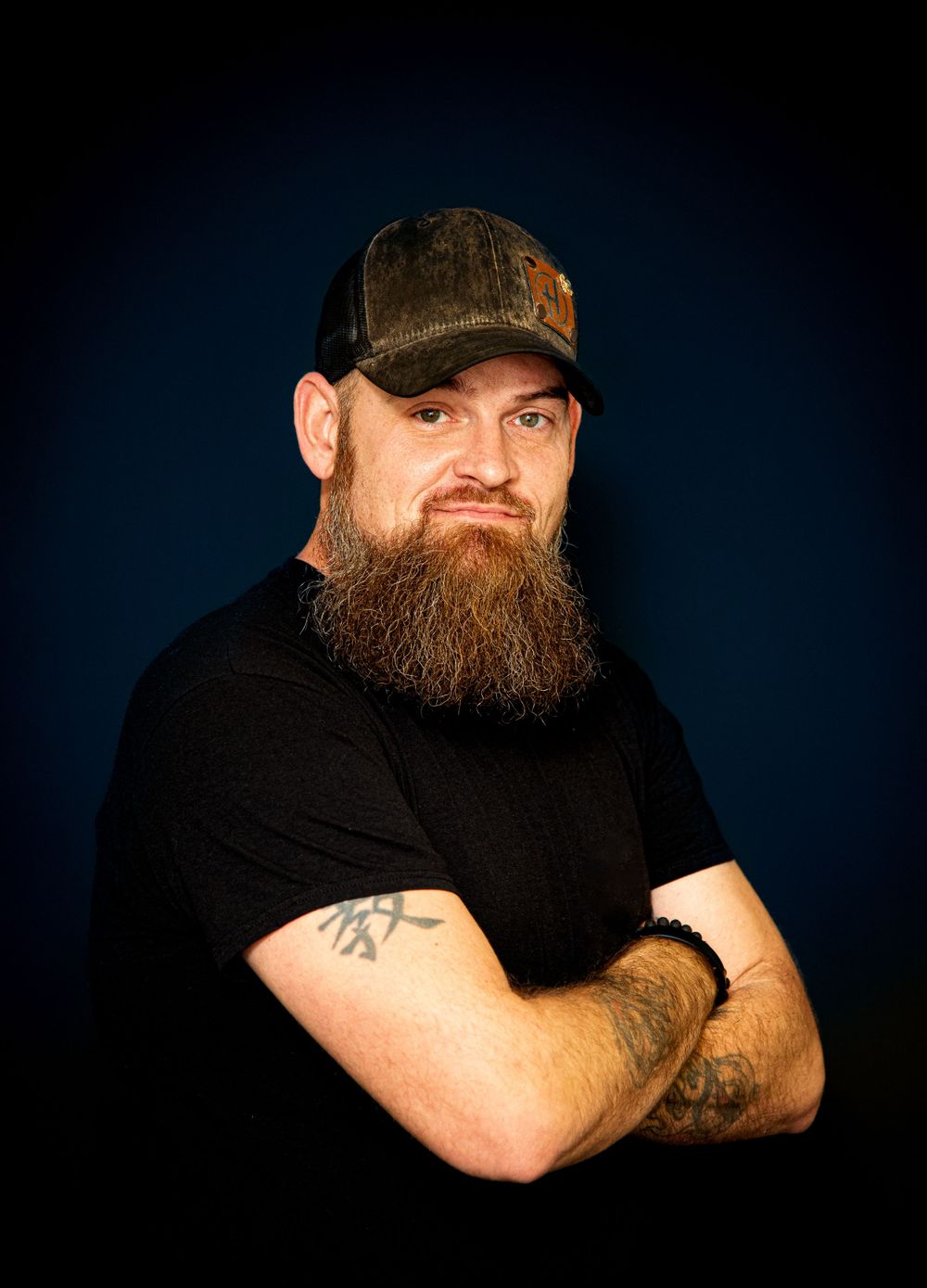 Studio portrait of a bearded person wearing a black t-shirt and baseball cap against a dark blue background.