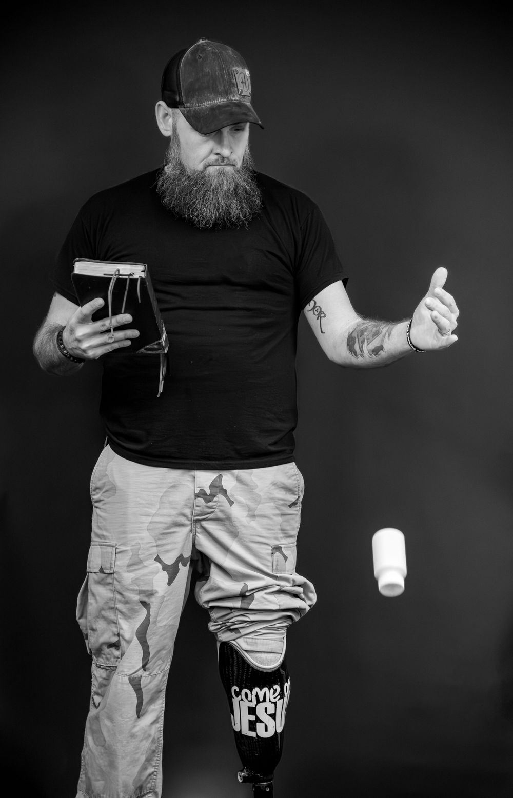 Black and white photo of person juggling a beer glass and can while wearing casual attire with tattoos.