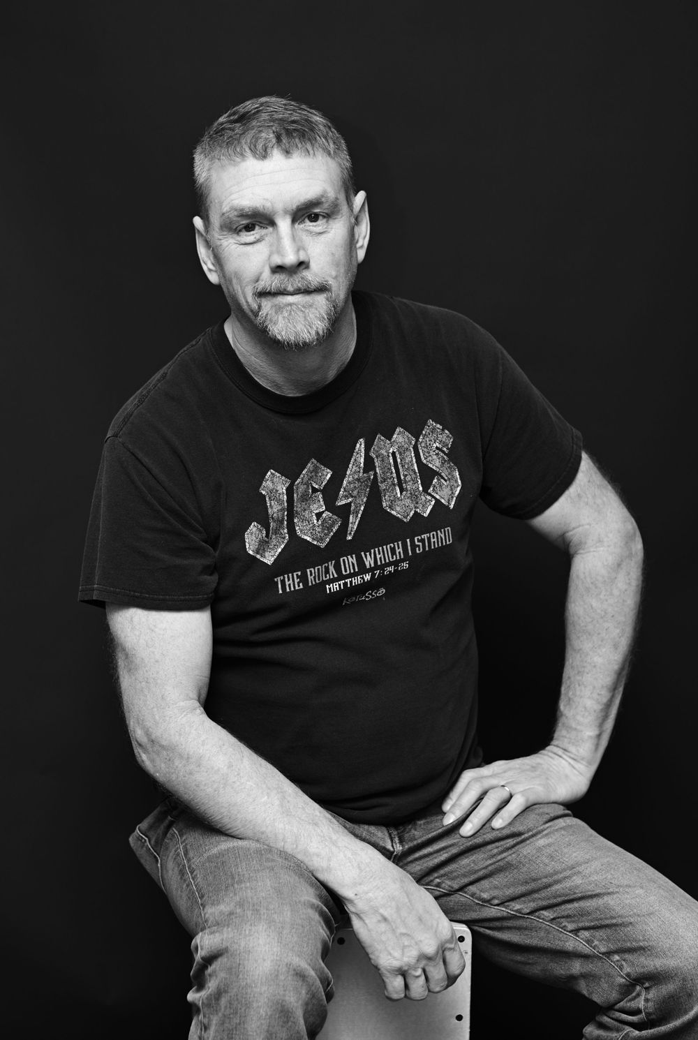 Black and white portrait of a person in an AC/DC t-shirt sitting casually against dark background.