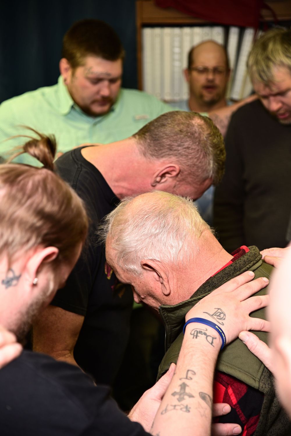 A group of people huddle together in prayer or meditation, with hands placed on each other's shoulders and backs.