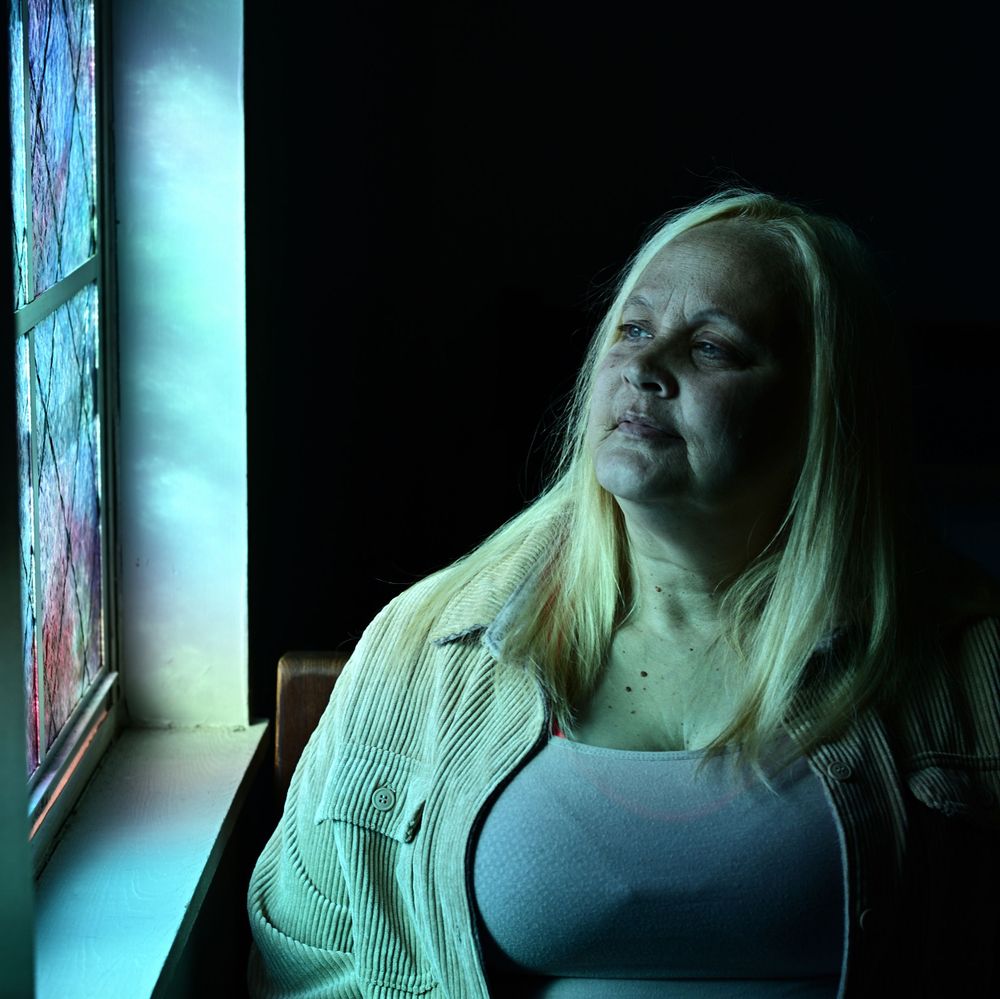 A person with long blonde hair gazes contemplatively through a window in dramatic blue lighting.