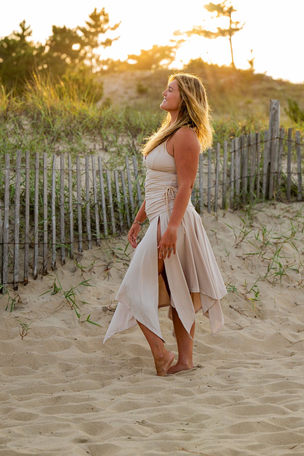 Figure in a flowing cream dress poses barefoot on sandy beach dunes at golden sunset with wooden fence in background.