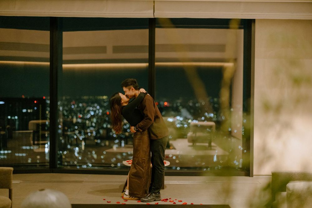 A couple embraces in silhouette against large windows overlooking a city skyline at night.