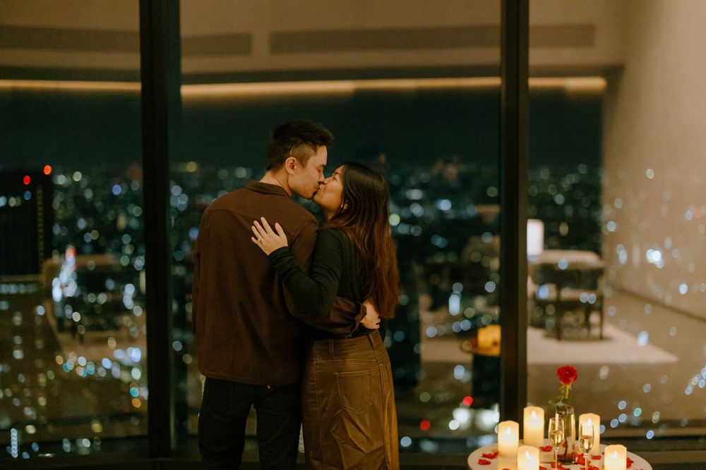 A couple shares a romantic moment by candlelit window with glittering city lights in the background.