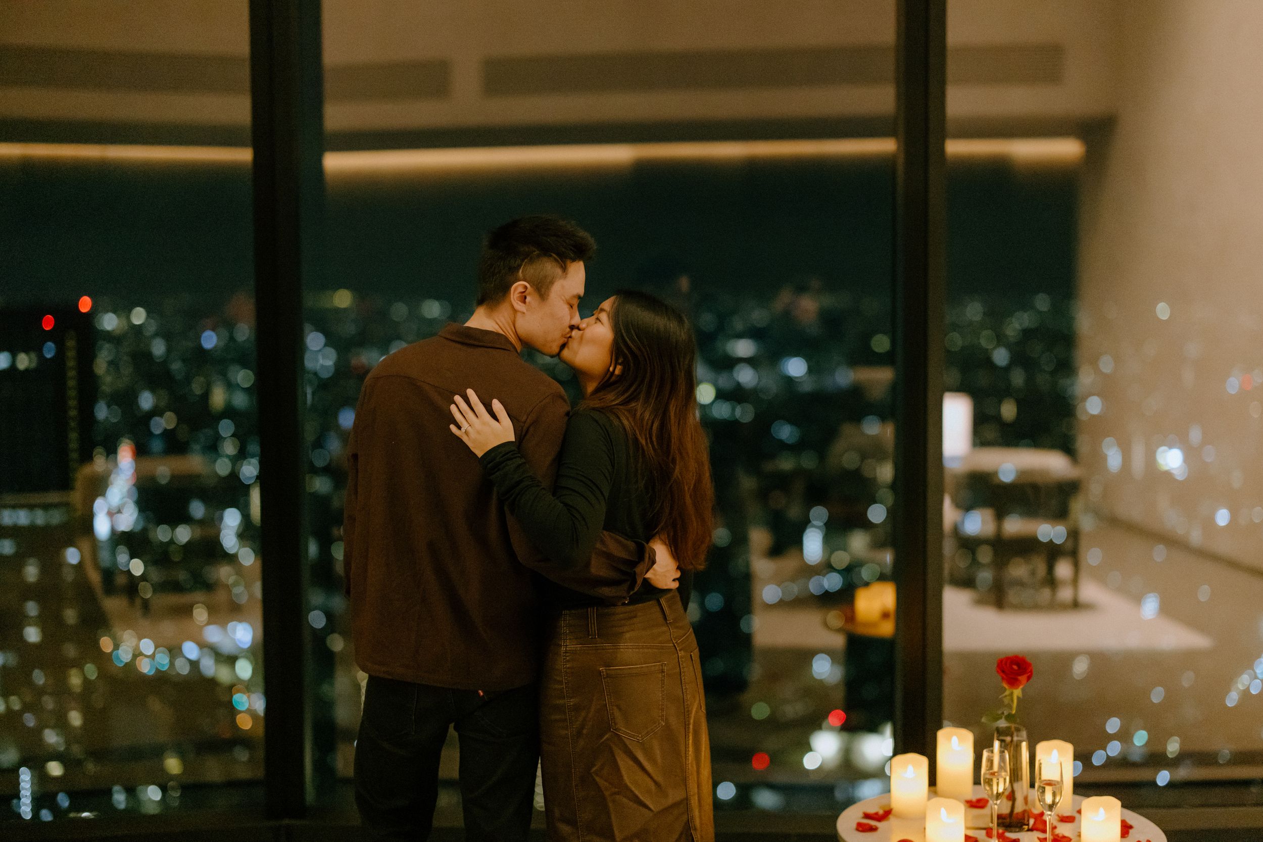 A couple shares a romantic moment by candlelit window with glittering city lights in the background.