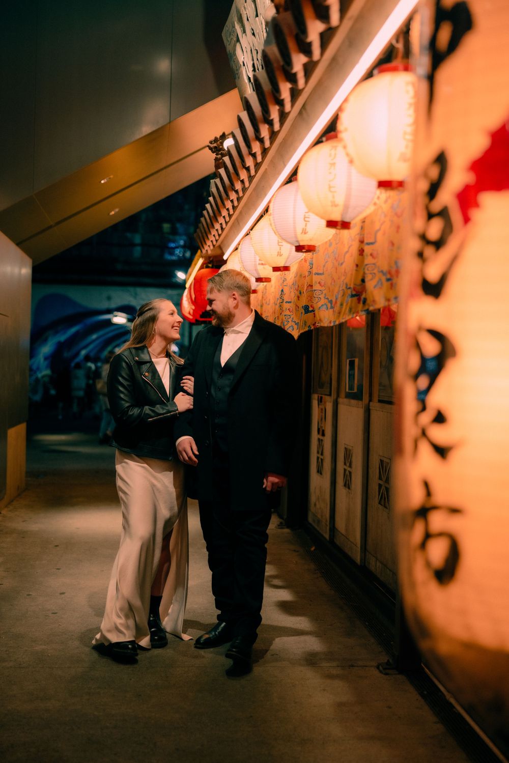 A couple shares an intimate moment in a dimly lit hallway decorated with glowing Asian-style lanterns and calligraphy.