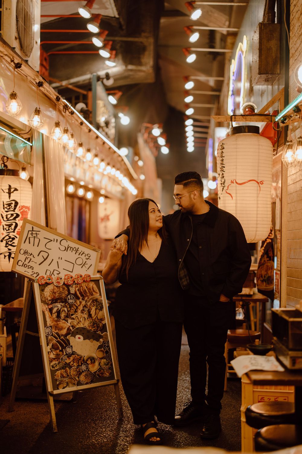 A couple stands in a cozy illuminated Japanese alleyway with string lights and restaurant signs at night.