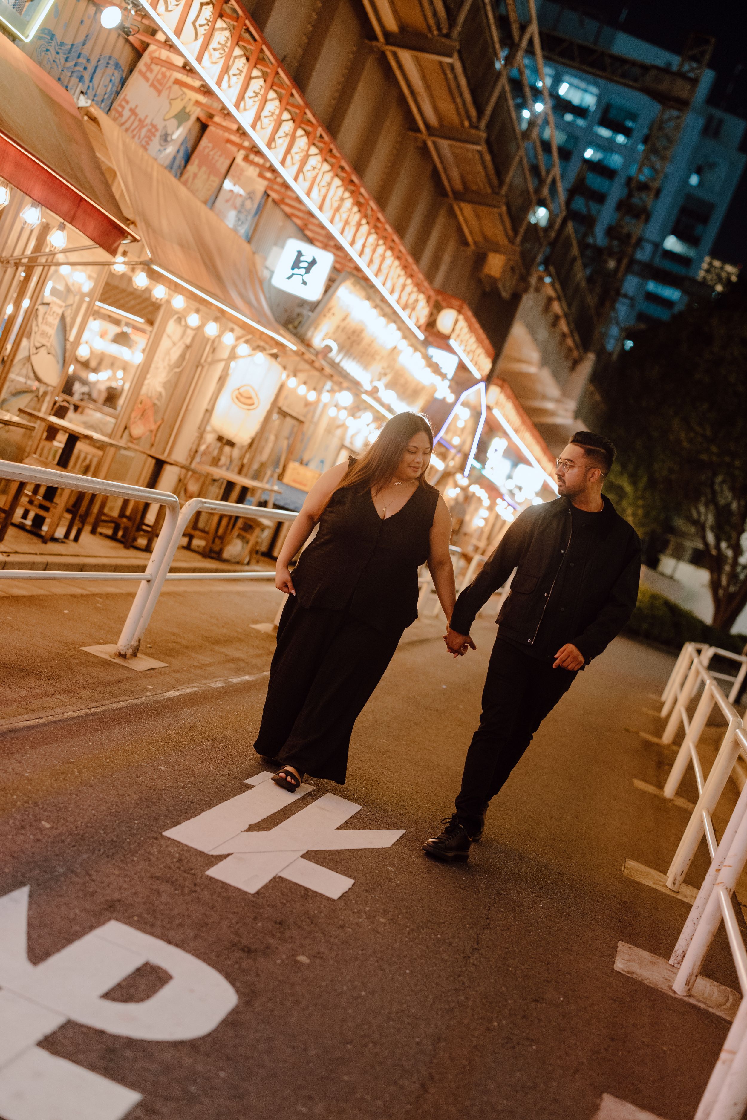 Couple holding hands walking on illuminated street at night with bright storefront lights in Japan.