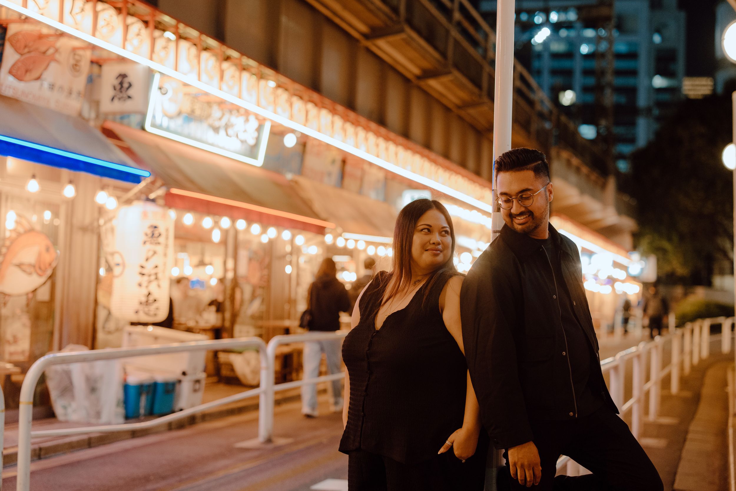 Young couple walking together at night past illuminated storefronts with warm lighting in an urban setting.