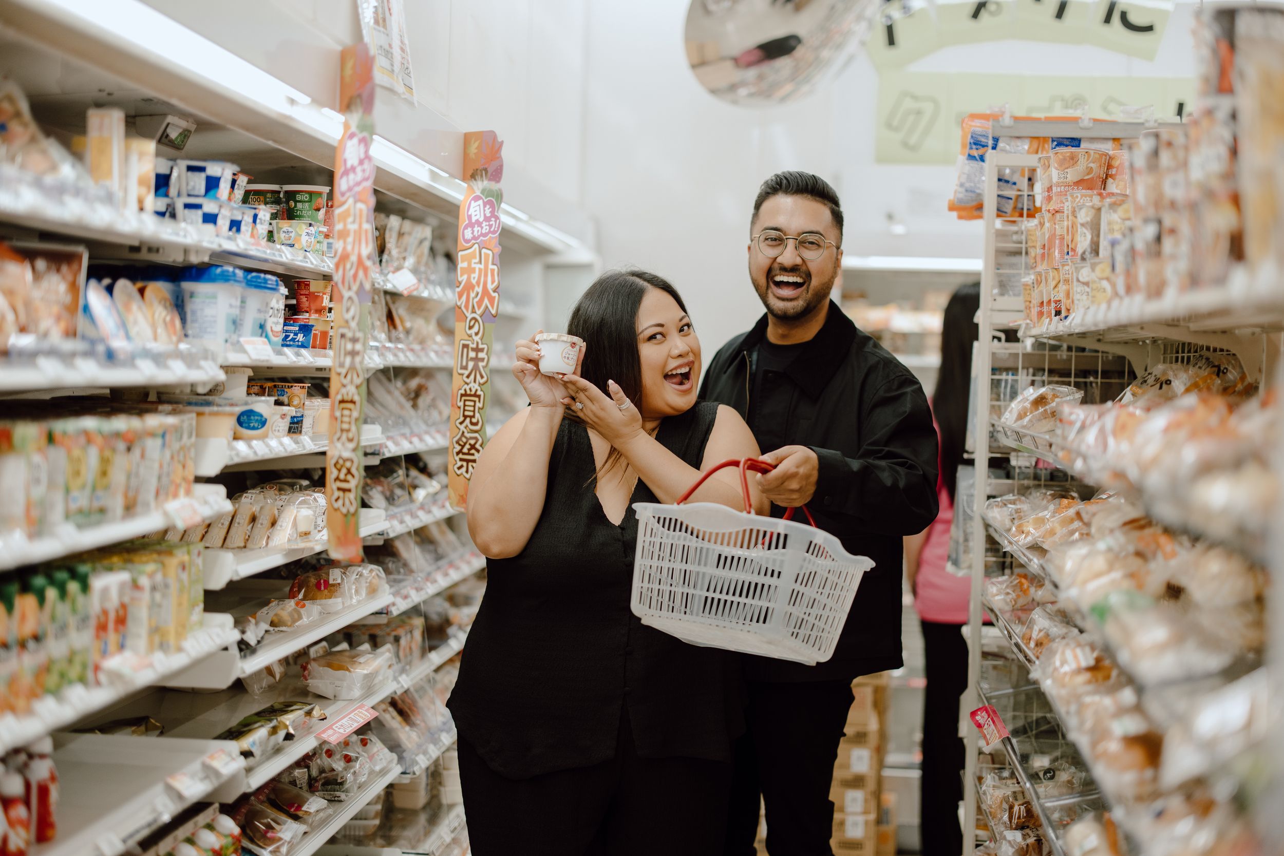 A couple laughs together while grocery shopping in a supermarket aisle holding a shopping basket.