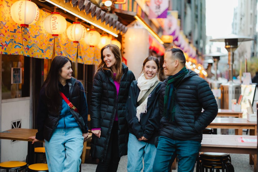 People dressed in winter coats chat and laugh together outside an Asian restaurant with paper lanterns in background.
