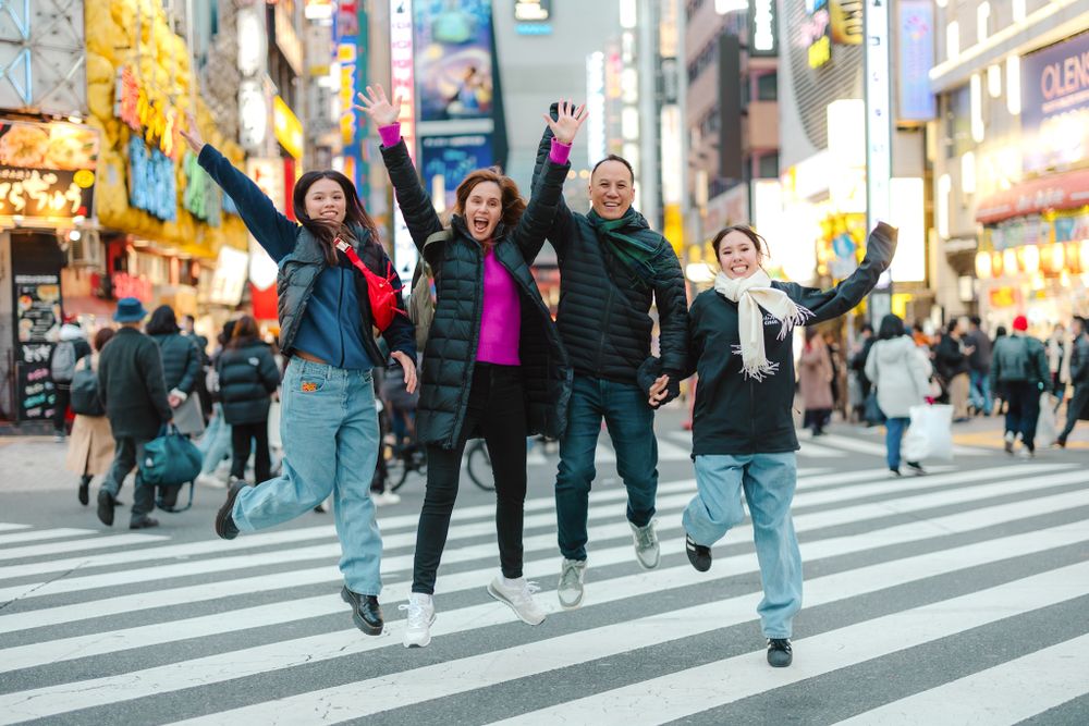 A group of friends jumping joyfully at a busy crosswalk in a vibrant Asian city street with colorful signage.