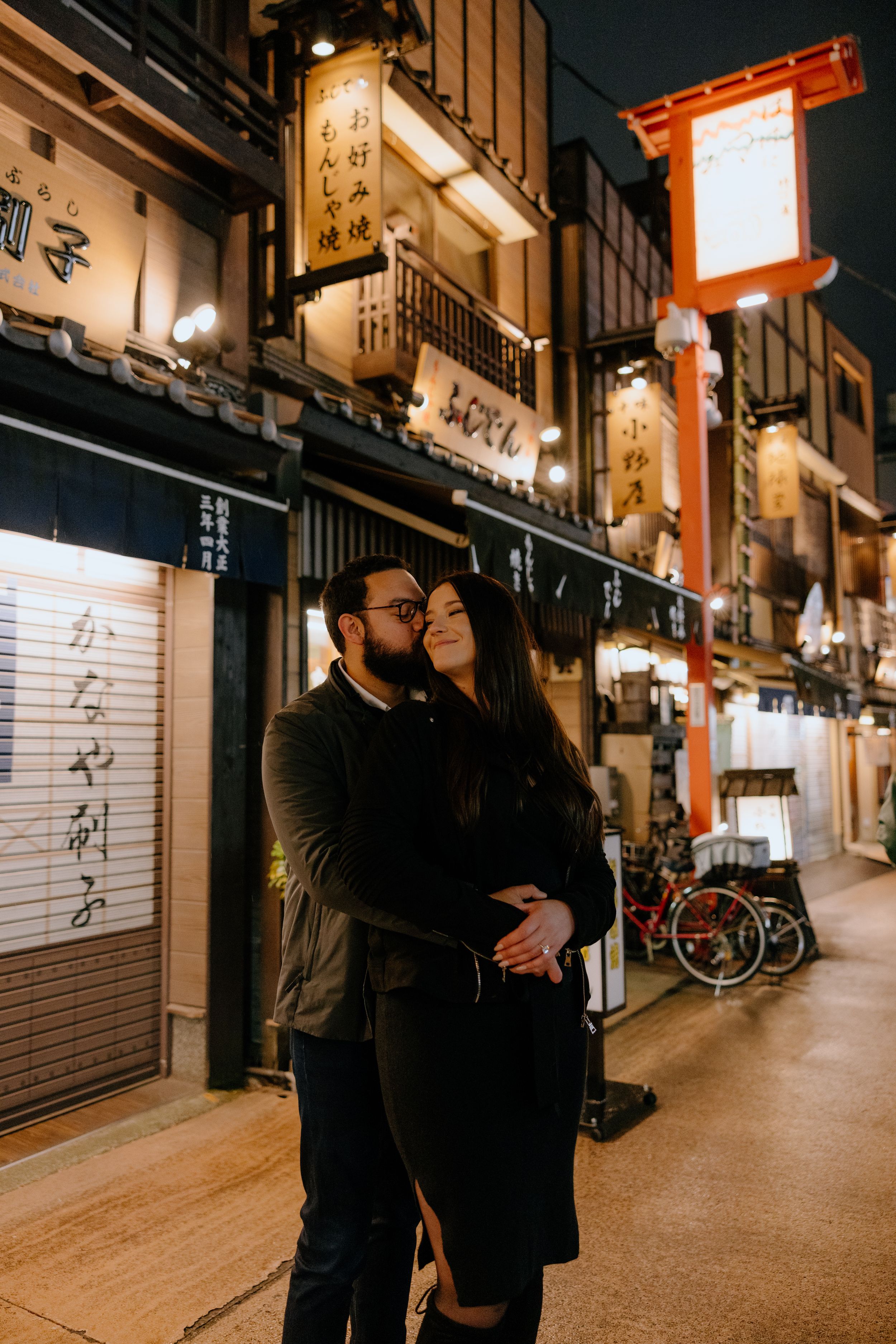 A couple embraces on a nighttime street in Japan with illuminated traditional lanterns and storefronts in the background.
