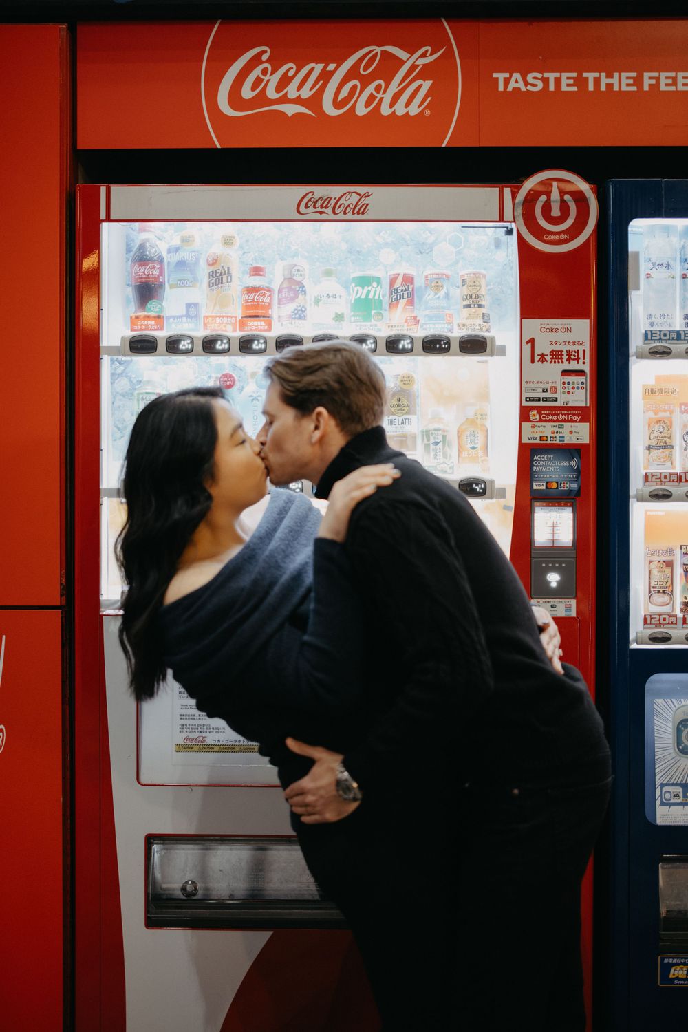 A romantic couple sharing a kiss in front of a red Coca-Cola vending machine at night.