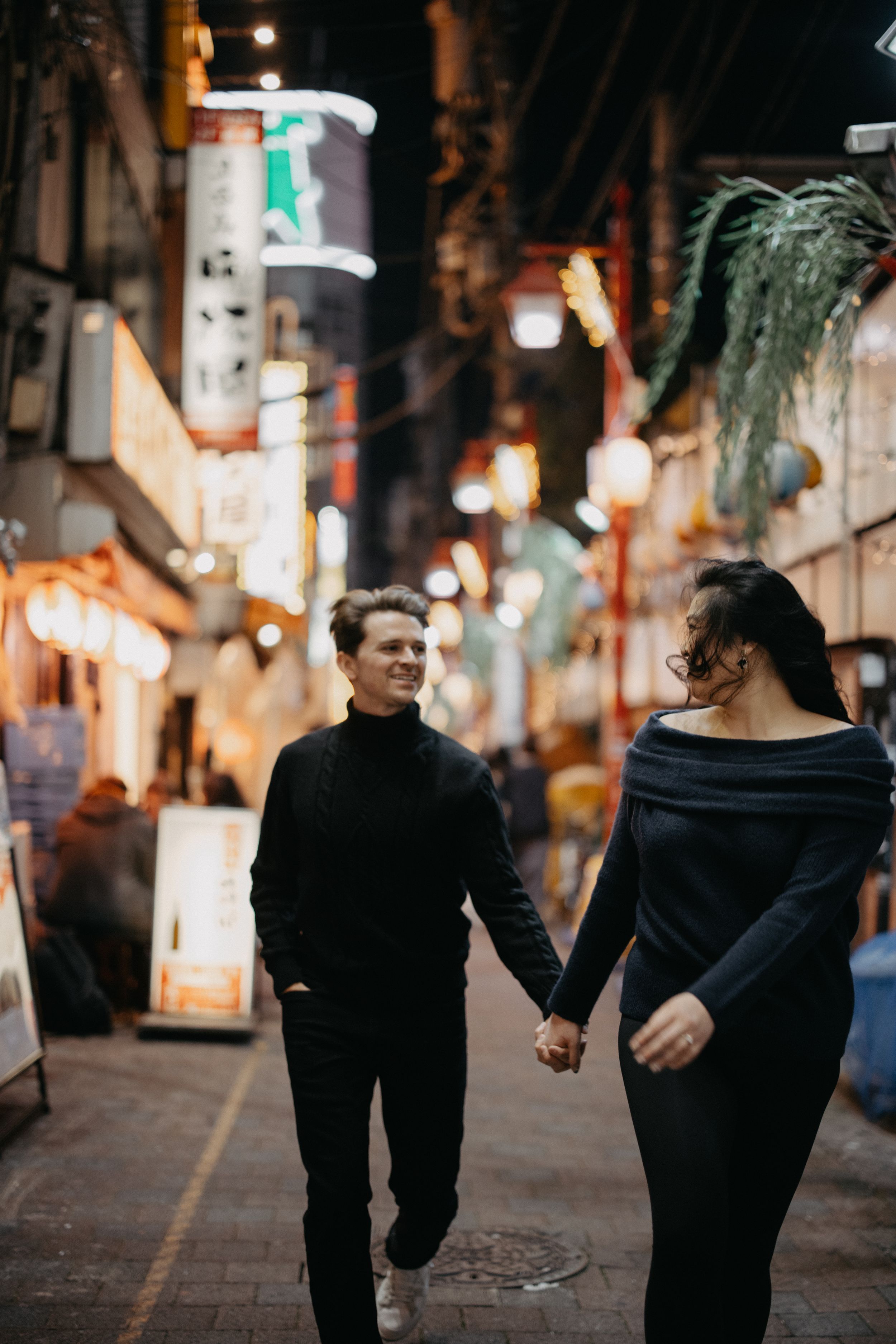 A couple walks hand in hand down a cozy urban street at night illuminated by warm storefront lights.