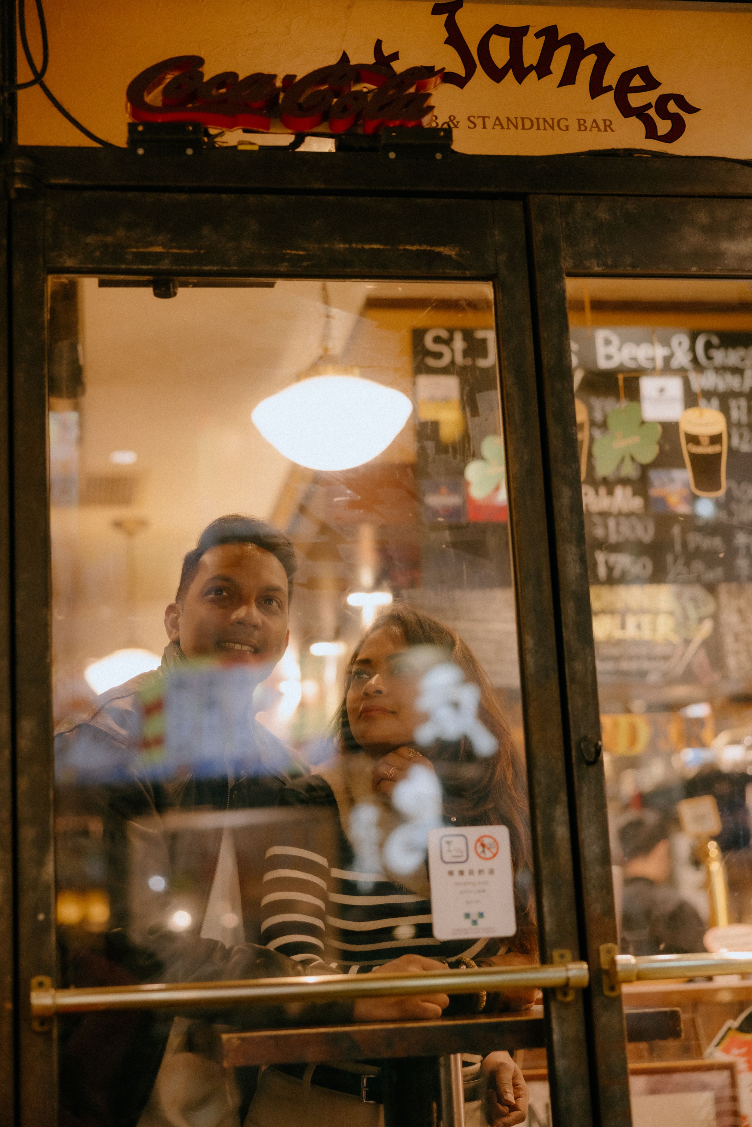 People peering through a glass window at Cheeky Jame's restaurant with warm interior lighting and reflections.