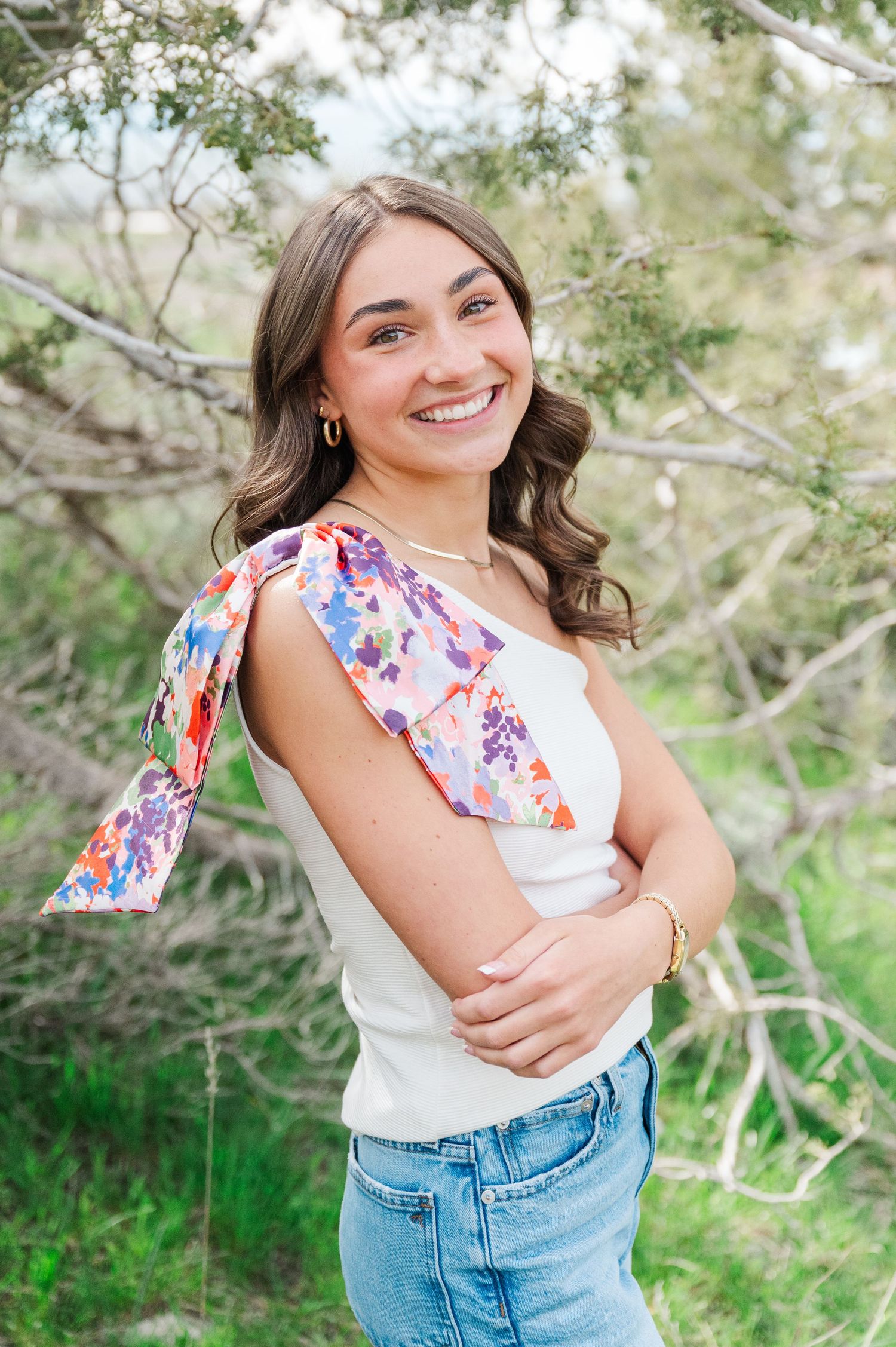 Person wearing a white top with floral accent and jeans poses outdoors against natural foliage background.