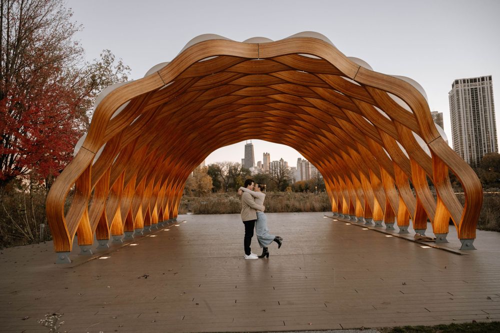 A couple stands beneath a wooden wave-like tunnel structure in a city park with fall foliage and skyscrapers in the background.