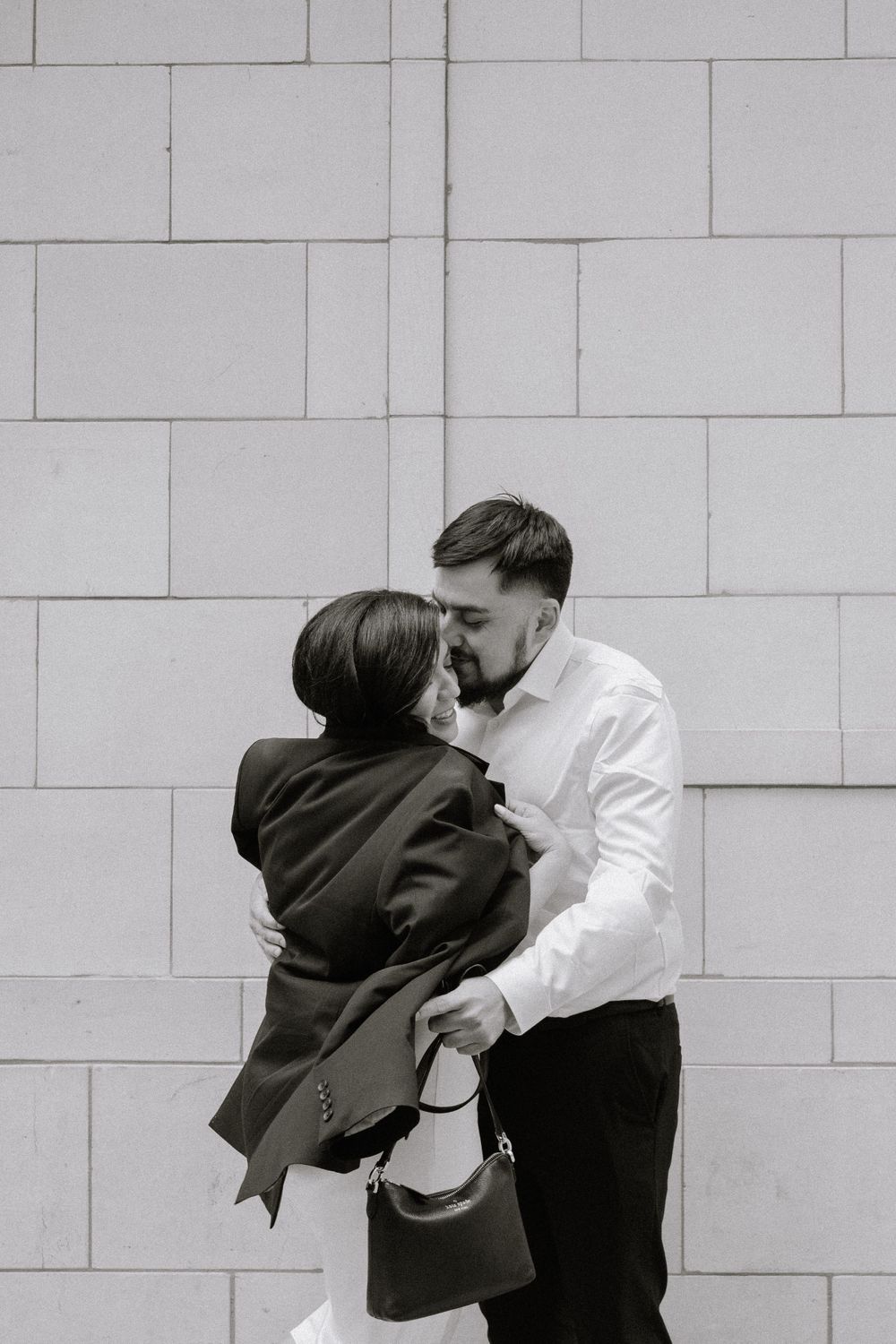 A romantic black and white portrait of a couple embracing against a modern concrete wall.