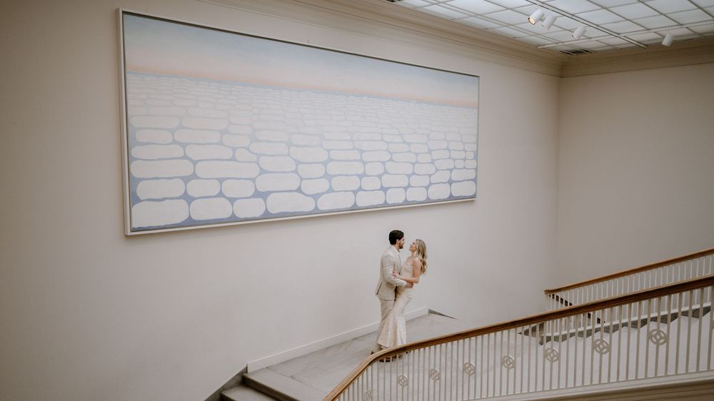 Bride and groom embracing on elegant staircase beneath large white abstract painting in art gallery wedding venue.